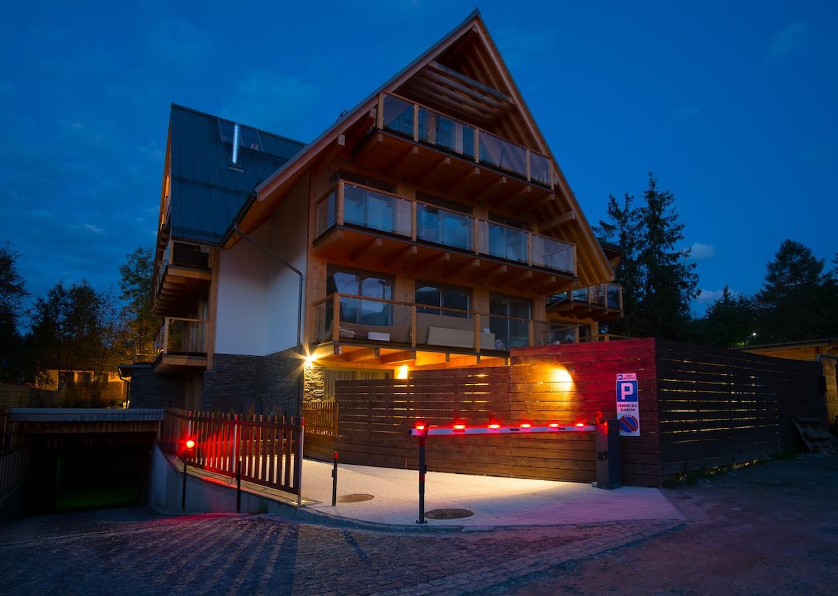 The exterior of the building is showcased at night, featuring wooden balconies and a sloped roof. Illuminated parking area is visible with a barrier indicating secure access to underground parking. Surrounding greenery adds to the tranquil atmosphere.