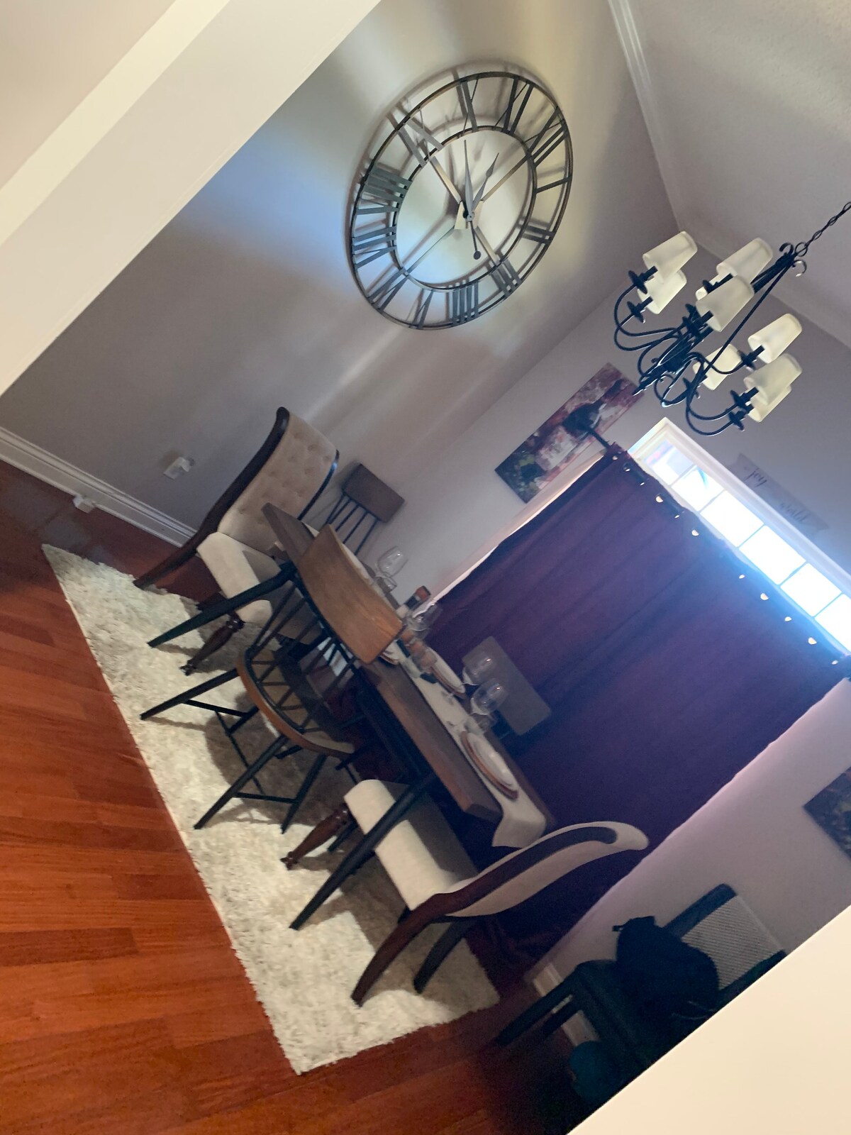 A dining area is shown with a wooden table surrounded by six chairs. A large wall clock hangs above the table, and a chandelier provides illumination. The space is accented by a fluffy area rug and dark curtains covering a window.