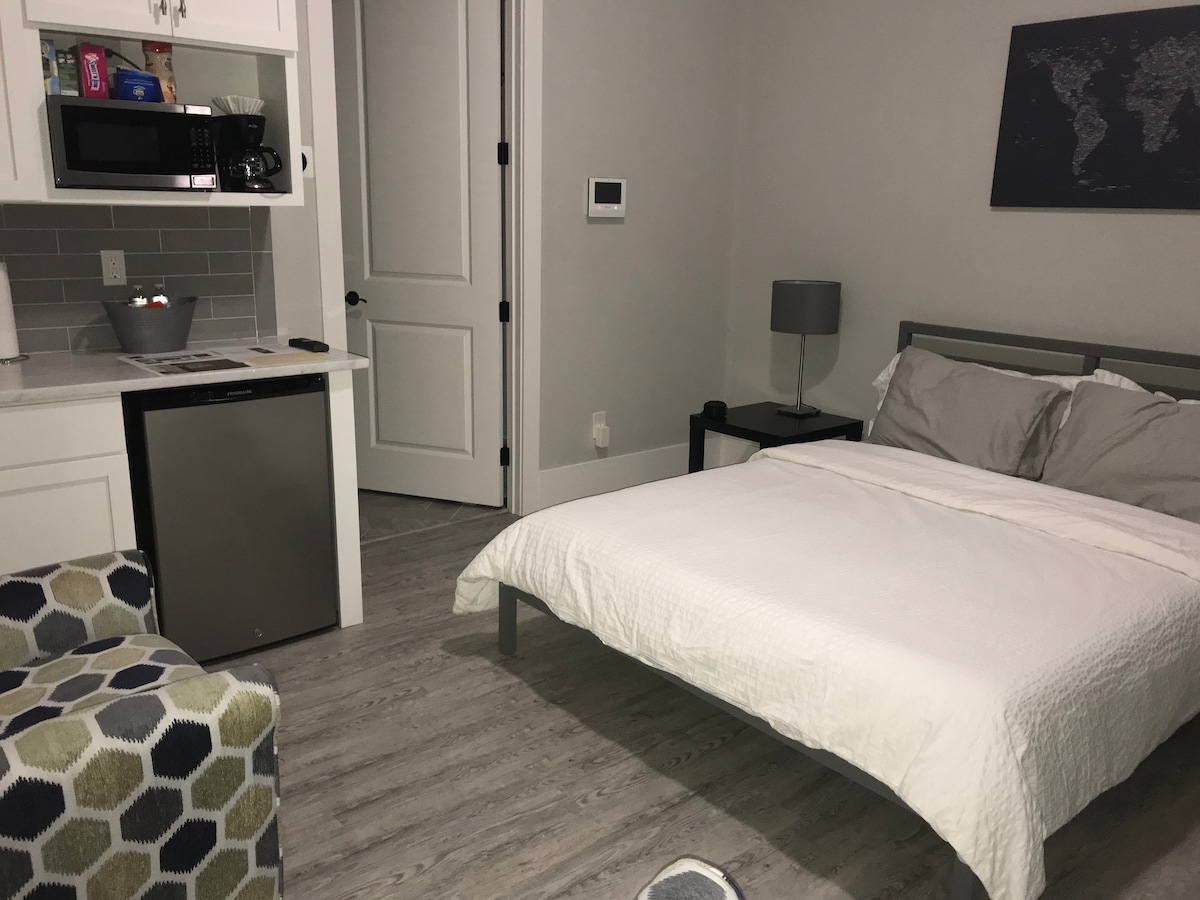 The in-law suite features a spacious bed with fresh white linens, complemented by a cozy armchair with a geometric pattern. A compact kitchen area includes a microwave and small refrigerator. Natural light floods the room through the nearby doorway, accentuating the calming color scheme.