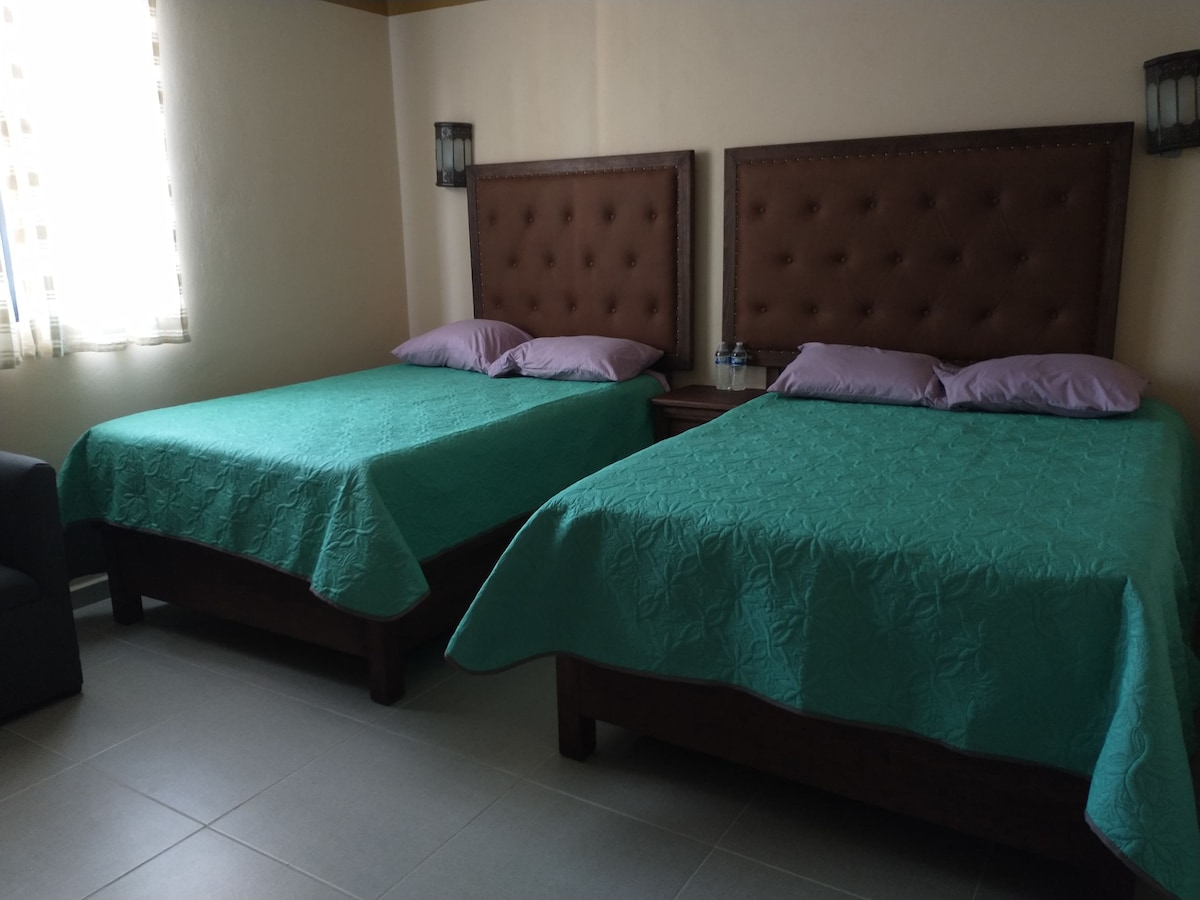 A room features two spacious beds, each adorned with teal covers and decorative purple pillows. A small seating area is visible in the corner, and natural light filters through sheer curtains, illuminating the neutral wall tones.