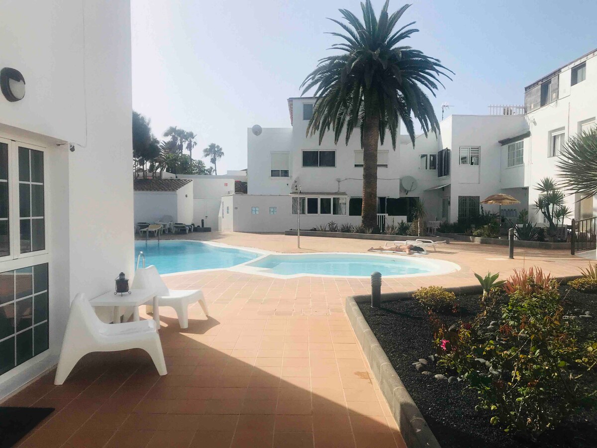 A tranquil outdoor space features a swimming pool surrounded by sun loungers and vibrant tropical plants. A large palm tree provides shade against a backdrop of white buildings, creating a serene environment for relaxation.