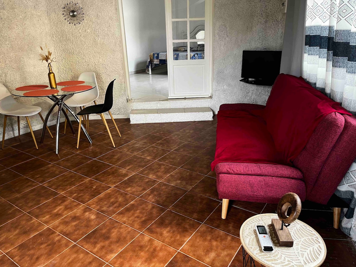 A comfortable living area features a red sofa and a circular dining table set with four chairs. A small side table holds decorative items. Light comes through a doorway leading to the sleeping area, while a television is mounted on the wall.