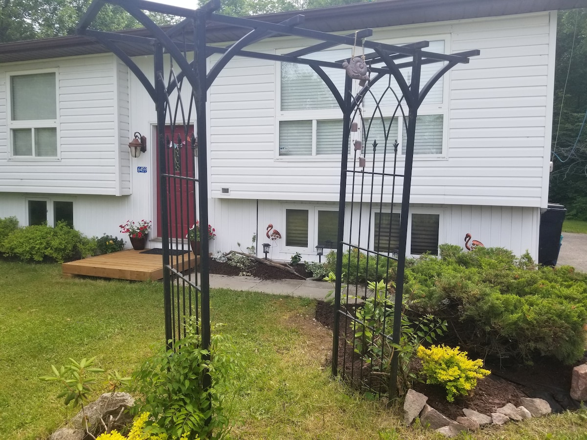 An inviting entrance is framed by a decorative archway, leading to a front porch with wooden steps. Lush green landscaping surrounds the entrance, including vibrant yellow flowers and neatly trimmed bushes. Two windows are visible, allowing natural light into the home.
