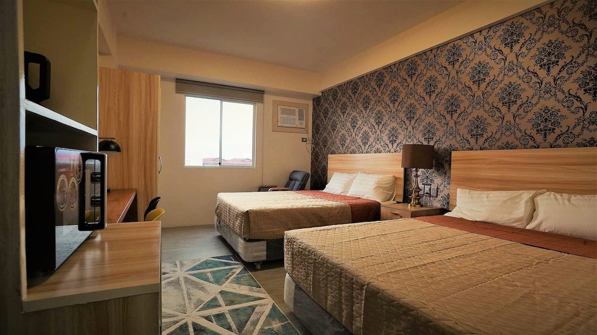 A spacious room features two large beds with warm-toned bedding. A patterned accent wall adds interest, while natural light enters through a large window. A small kitchenette is visible, alongside a comfortable seating area and a desk, creating a functional space for relaxation.