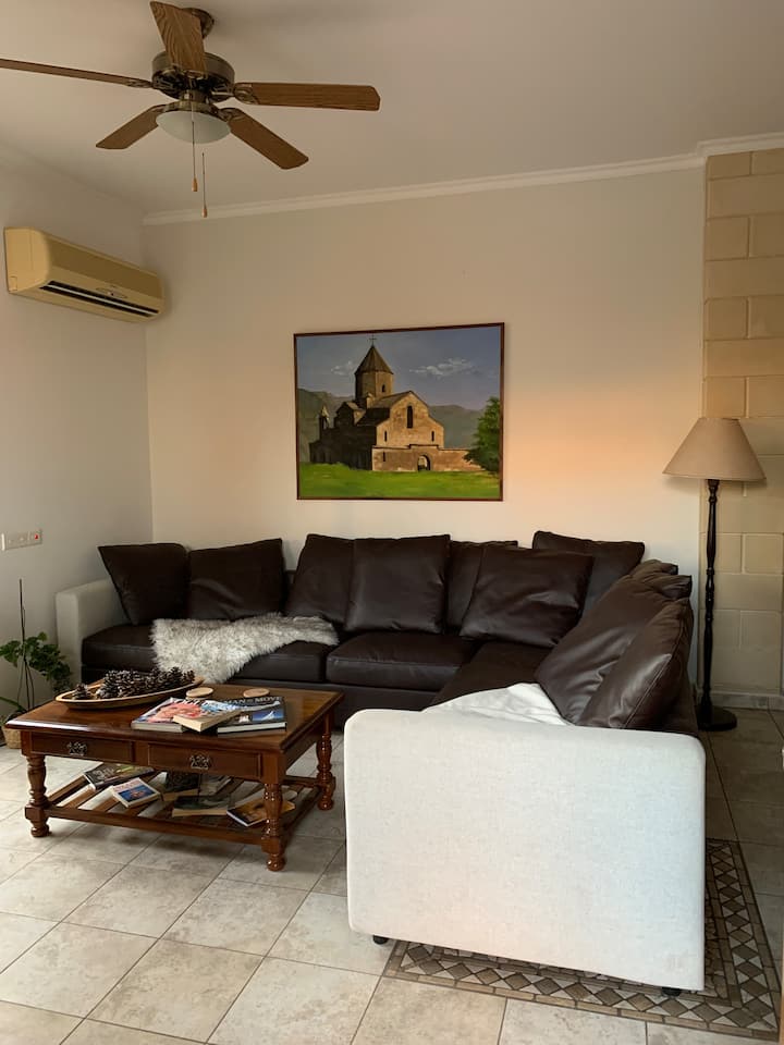 Всекидневна на открито с голям ъглов диван, масичка за кафе, телевизор и изглед към градината. За ваше удобство има вентилатори на тавана и климатик.