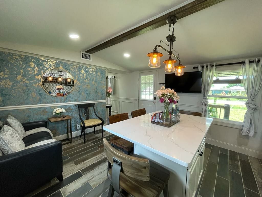 A modern dining area features a white marble island surrounded by wooden chairs. The room is accented by a floral wallpaper and a decorative mirror. Natural light streams in through the window, illuminating the cozy seating area with a dark sofa and decorative pillows.