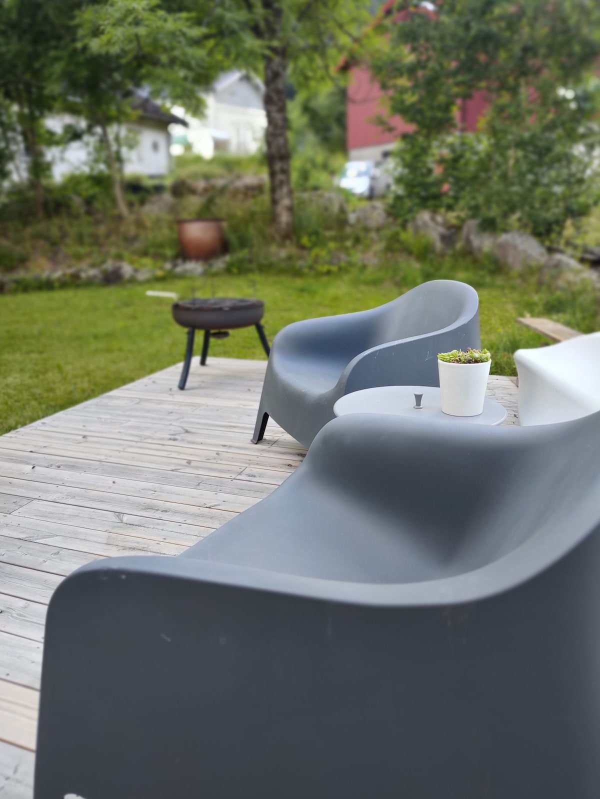 An outdoor seating area is depicted on a wooden deck, featuring two modern, curved chairs in shades of gray. A small table with a potted plant occupies the center space, accompanied by a grilling area in the background. Lush greenery surrounds the setting.