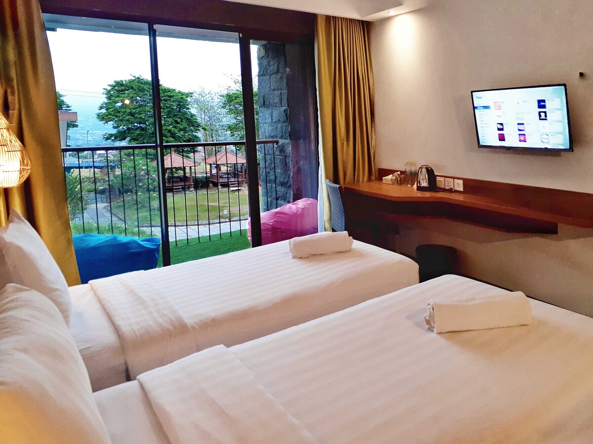 A well-lit guestroom features two beds with neatly arranged white linens. A balcony with sliding glass doors offers a view of green outdoor spaces. A television is mounted on the wall, while a small countertop displays essentials.