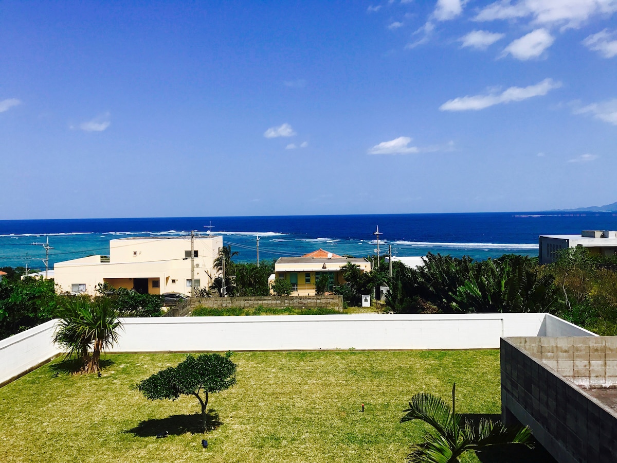A view of the eastern coast with the East China Sea extending to the horizon. A lush green lawn is visible in the foreground, contrasting with the blue ocean waters. Several houses can be seen in the background amidst tropical vegetation under a clear sky.