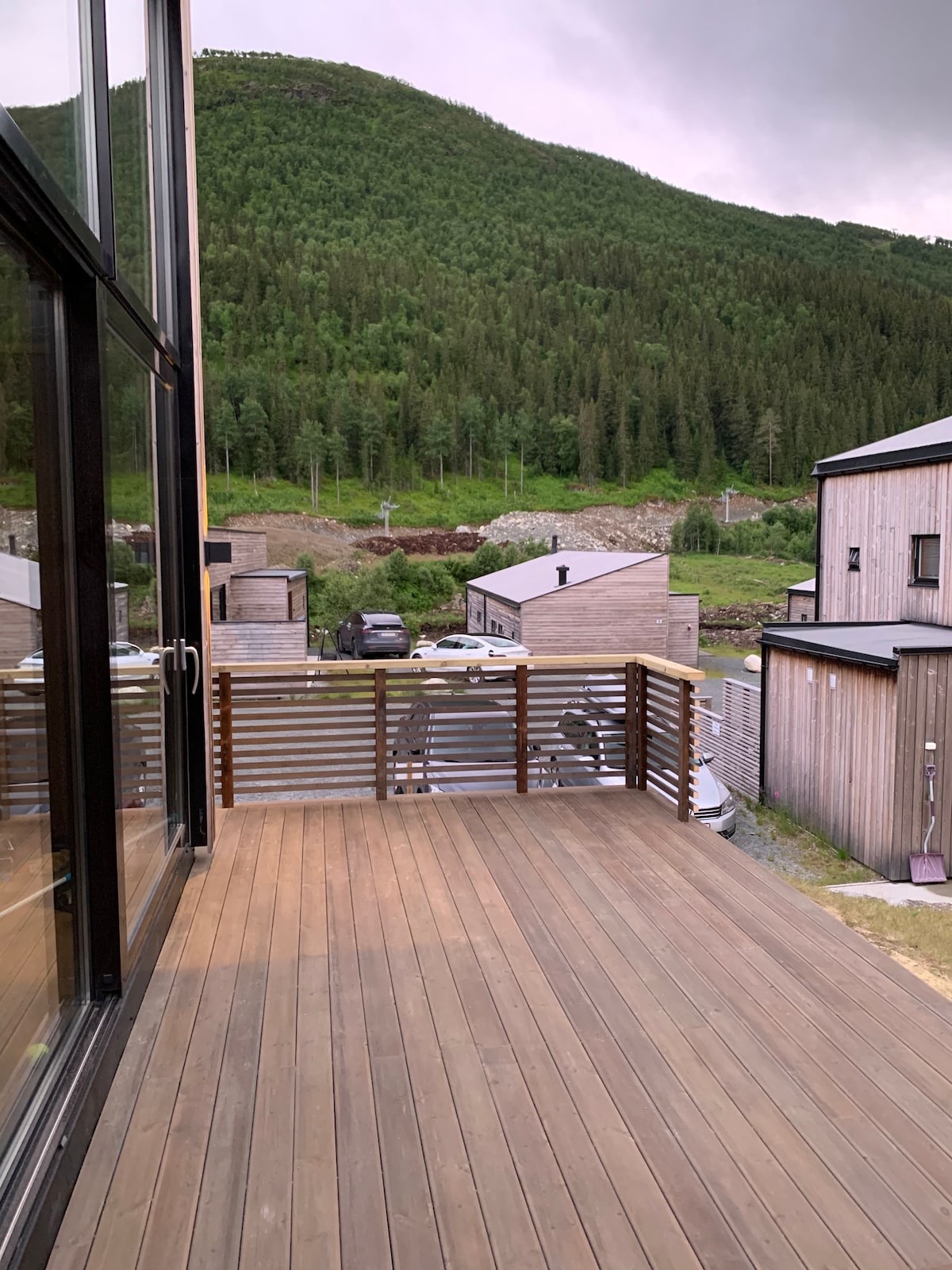 A wooden deck extends from the modern cabin, offering views of the surrounding hillside and homes. The area is characterized by its natural greenery and wooden structures, with vehicles parked nearby, blending the outdoor space with the serene landscape.