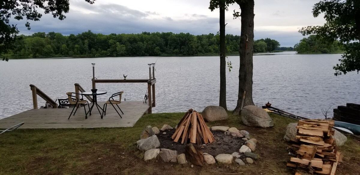 A serene riverside patio features a wooden deck with two chairs and a small table. Nearby, a stone fire pit holds neatly stacked firewood, surrounded by a landscaped area. The expansive waters of the St. Joe River reflect the overcast sky, bordered by lush green trees.
