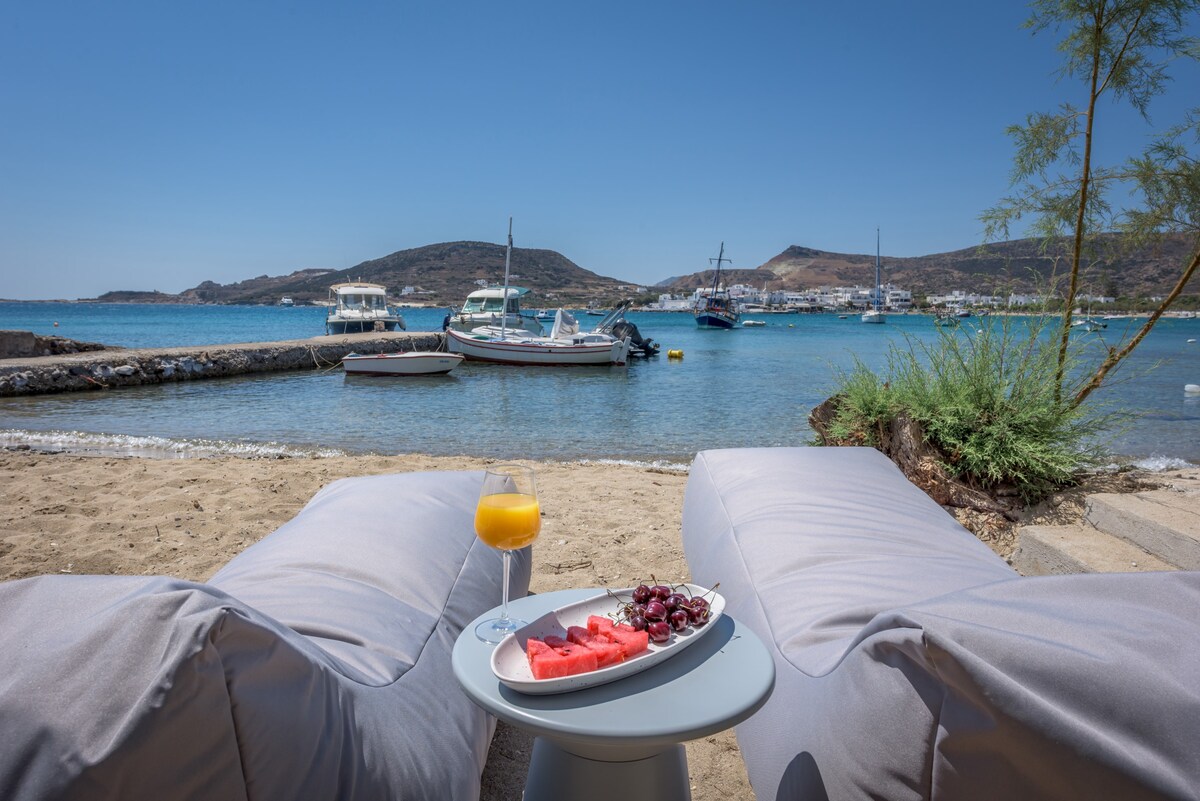 Two comfortable loungers are positioned on the sandy beachfront, offering a view of the clear blue water and moored boats. A small table holds a plate of fresh fruit and a glass of orange juice, enhancing the serene seaside atmosphere.