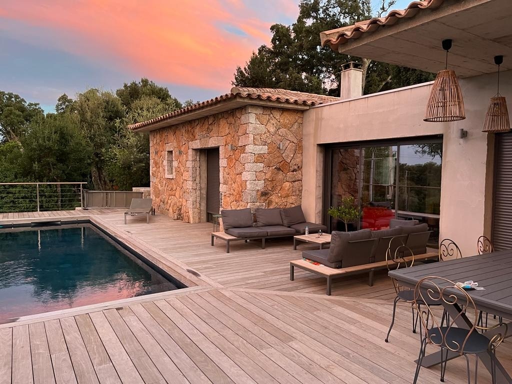 The exterior view showcases a charming stone house with a modern design. A wooden terrace surrounds a tranquil swimming pool, complemented by an outdoor seating area featuring comfortable sofas. A dining table, situated nearby, invites leisurely meals al fresco under the changing sky.