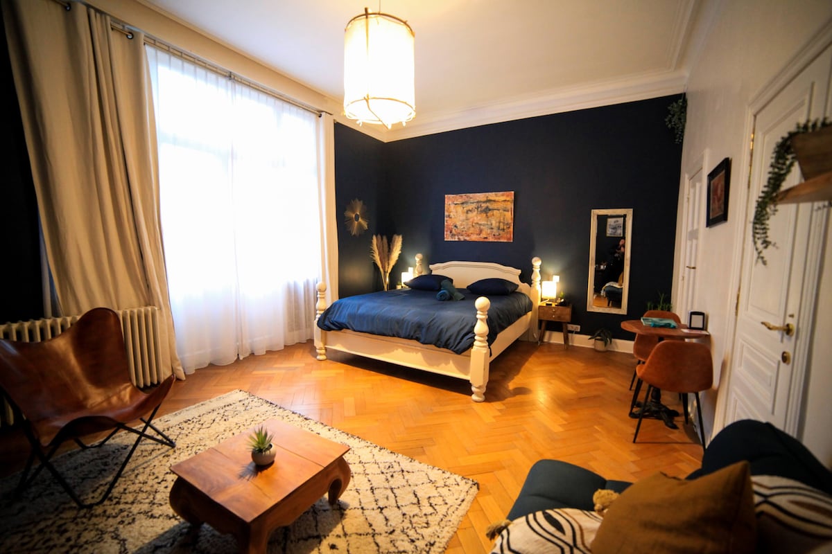 A spacious bedroom features a large white bed dressed in navy linens. Natural light streams in through tall windows, illuminating the dark blue accent wall. A cozy seating area is arranged beside a small wooden table, with a unique texture rug enhancing the room's warmth.