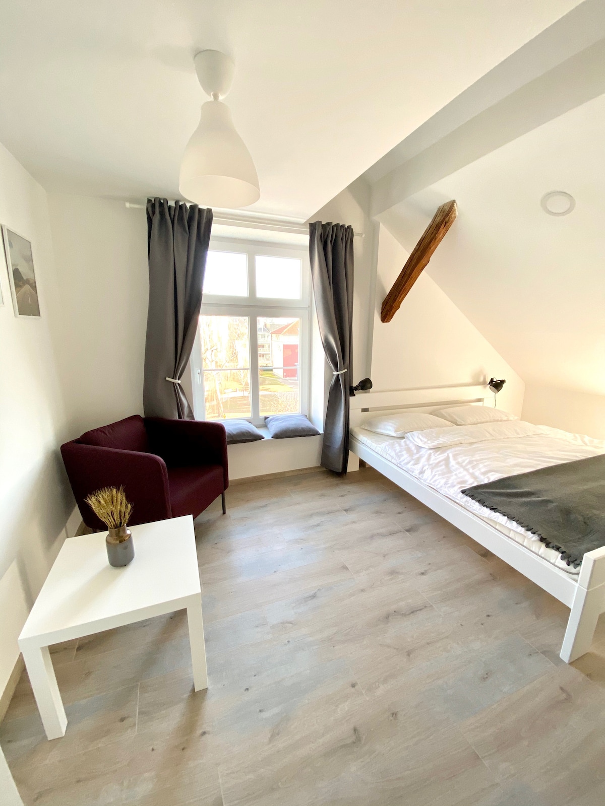 The room features a queen bed dressed in crisp linen, positioned near a bright window with gray curtains. A cozy reading nook is created by a red armchair beside a small white table. Natural light enhances the simplicity of the space with wooden flooring.