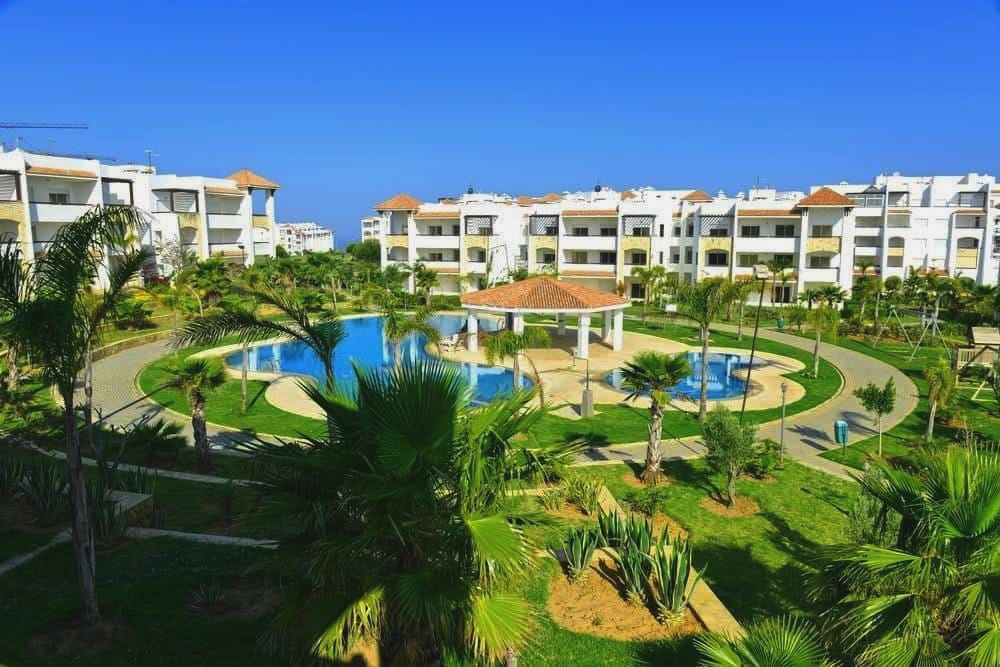 A lush landscape features well-manicured gardens with palm trees surrounding several swimming pools. The bright blue sky complements the white residential buildings in the background, emphasizing a serene atmosphere. Pathways meander through the green spaces, leading to various recreational areas.