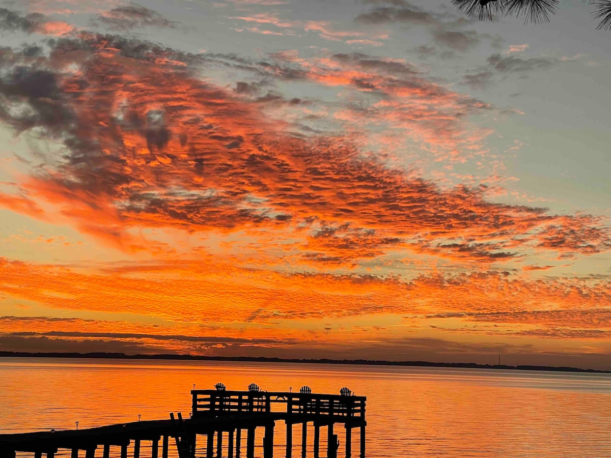 A vibrant sunset casts a warm glow over the water, reflecting shades of orange and pink. Silhouetted against the colorful sky, a rustic pier extends into the calm water, creating a tranquil scene.