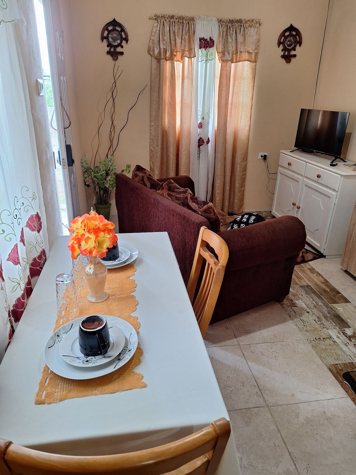A dining area is set with two plates and cups on a white table, adorned with an orange runner. Nearby, a cozy seating arrangement features a brown sofa and a small TV on a white cabinet, with light filtering through frosted curtains.