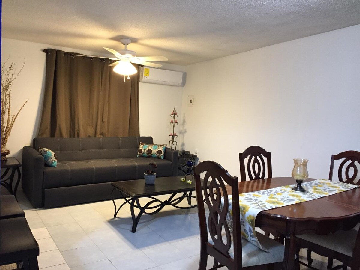 A living area features a dark gray sofa, complemented by decorative pillows, and a ceiling fan above. A wooden dining table with chairs is present, highlighted by a table runner. Light-colored tiles reflect natural light from the nearby window, enhancing the spacious feel.