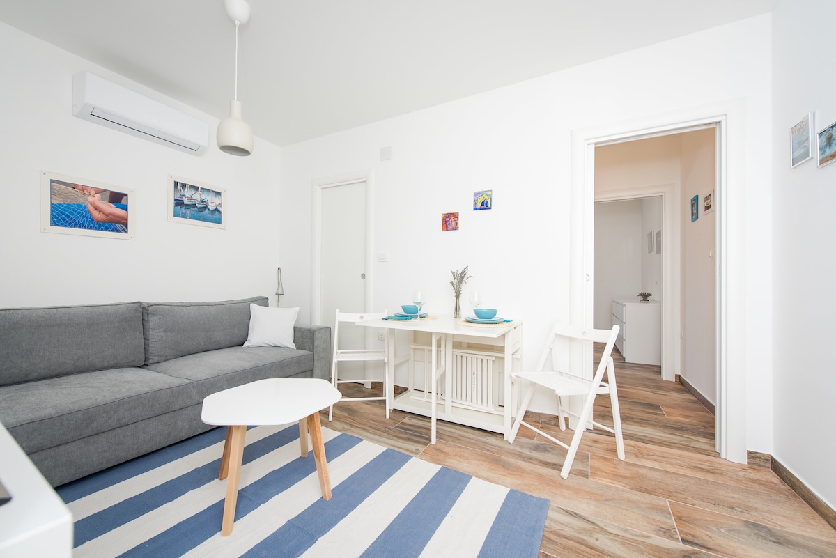 A modern living space features a gray sofa paired with a round coffee table set atop a striped area rug. A dining table with light-colored chairs is positioned nearby, with colorful photographs adorning the walls. Bright light fills the room through the doorways.