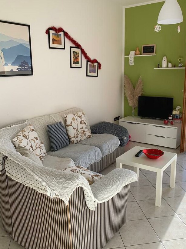 A living area features a striped couch adorned with patterned cushions and a cozy throw. A small white coffee table sits in front, surrounded by artwork on the walls. A flat-screen television is positioned on a low white unit, complemented by decorative items on a shelf.
