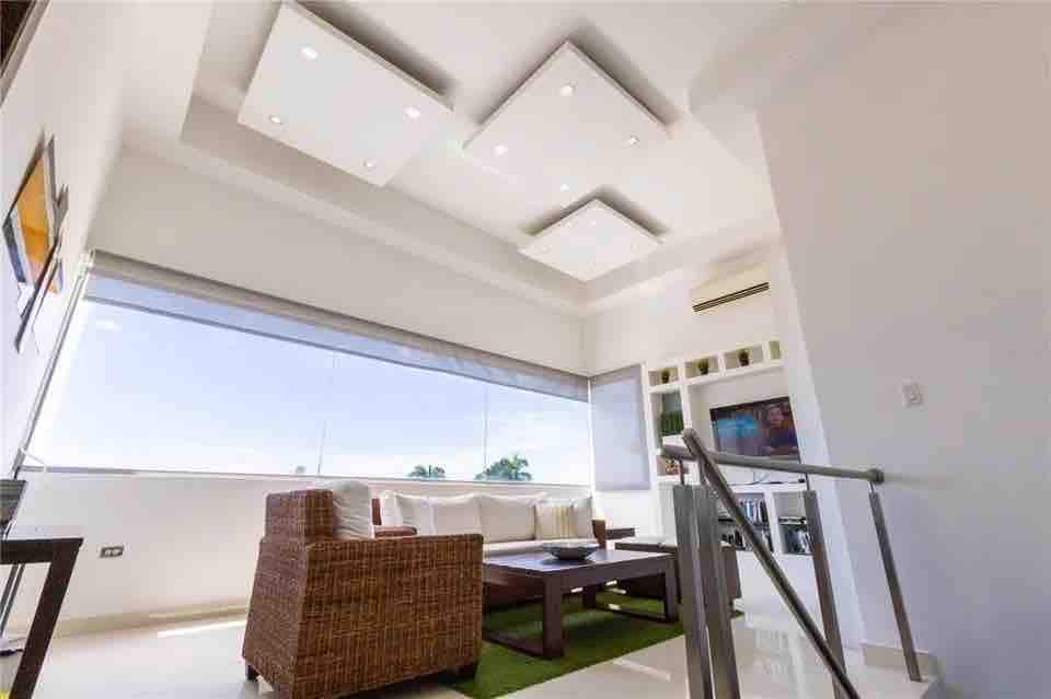 A modern living area features a large window allowing natural light to enhance the space. A comfortable seating arrangement is set atop a green area rug, with a wooden coffee table in the center. Sleek ceiling lights provide ambient illumination.