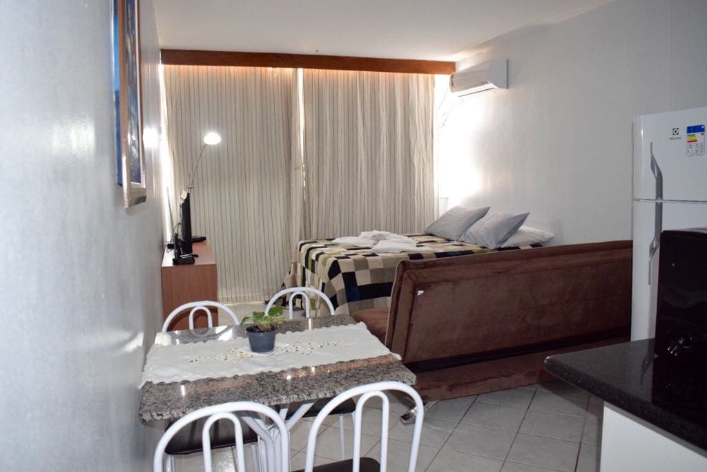 A cozy studio layout features a spacious sleeping area adjacent to a dining space. An air conditioning unit is visible above the bed, while bright natural light streams through large curtains. The dining table is set with four chairs, and a small television is mounted on the wall.