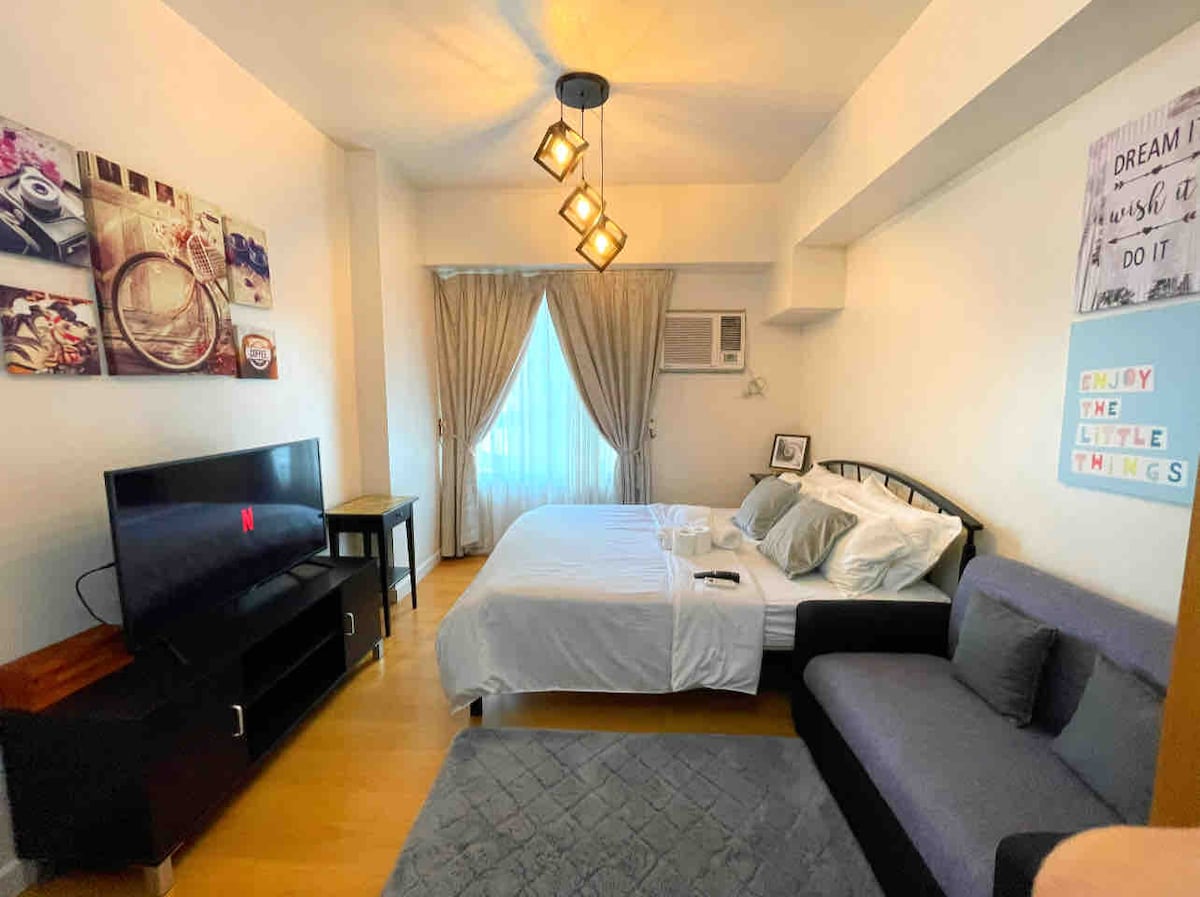 A cozy sleeping area features a spacious bed dressed in fresh linens, accompanied by a modern television on a low black console. A gray sofa provides additional seating. Natural light enters through the large window, which is framed by elegant drapes.