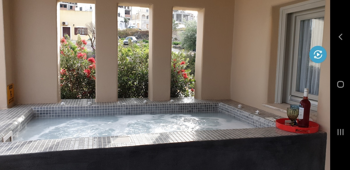 A private jacuzzi is surrounded by stone-tiled seating, situated under an open window with shrubby greenery visible outside. The bubbling water reflects a serene atmosphere, while a tray with a decorative piece sits at one end, enhancing the calming environment.