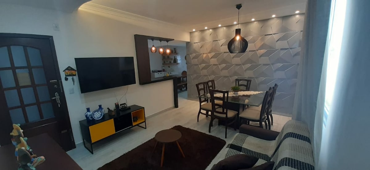 A stylish living area is presented with a modern dining table surrounded by chairs. A wall with a textured design serves as a backdrop, while a television is mounted on the opposite wall. Soft lighting fixtures provide an inviting ambiance.