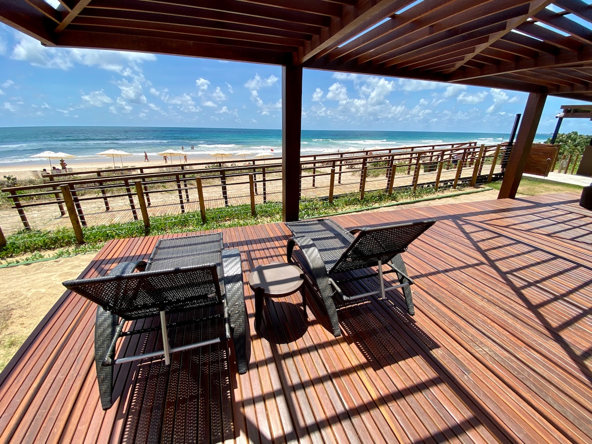 A spacious deck features two lounge chairs positioned for ocean views. The wooden flooring complements the surrounding landscape, while a railing offers a barrier leading to the sandy beach. Bright sunlight highlights the serene beachscape under a clear blue sky.