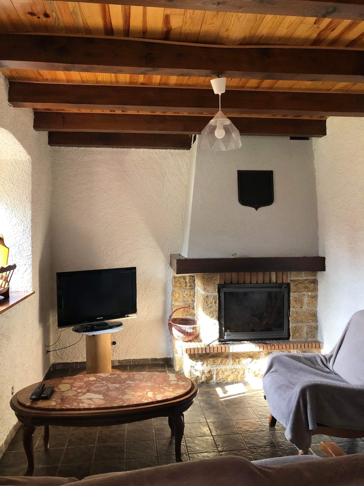 A cozy living area is featured with a stone fireplace and a television on a small stand. Natural light enters through a nearby window, illuminating the space. A simple table with a marble top rests beside a soft seating area, complemented by wooden beams overhead.