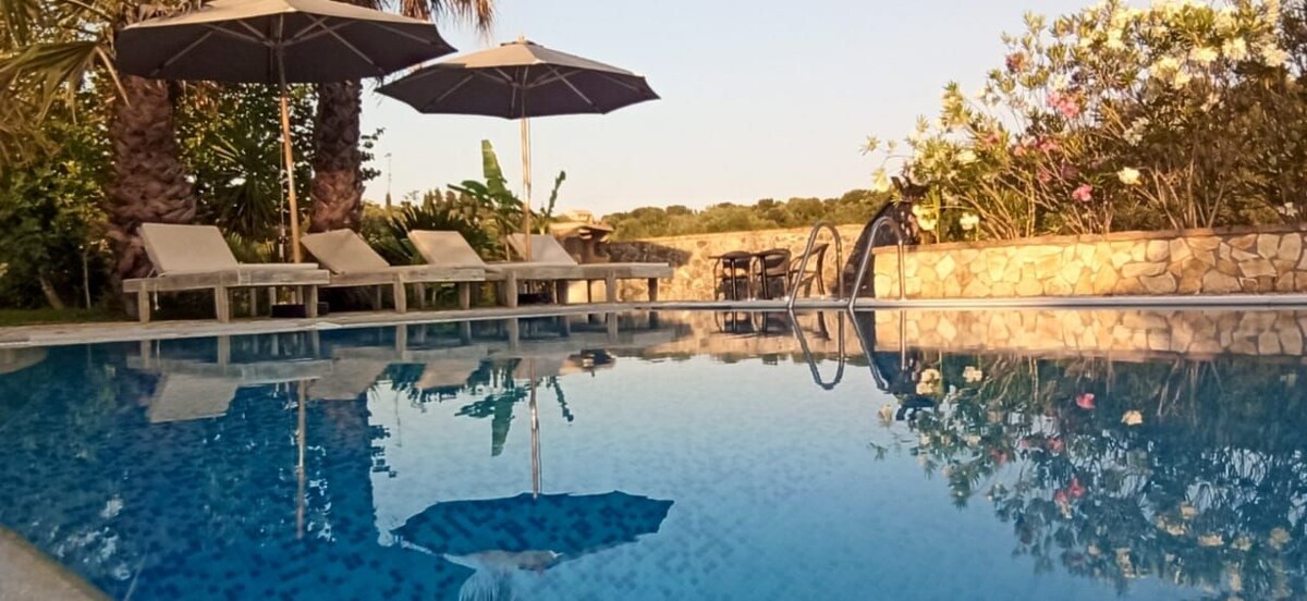 A tranquil swimming pool is surrounded by sun loungers with parasols, reflecting the surrounding greenery. The pool area is framed by low stone walls and features lush plants and palm trees, creating a serene outdoor environment.