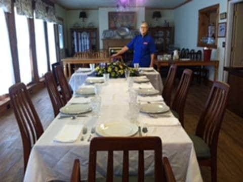 A spacious dining area is arranged with a long table set for a meal. White tablecloths cover the table, with place settings that include plates and glassware. Wooden chairs surround the table, and large windows allow natural light to fill the room, enhancing its welcoming feel.