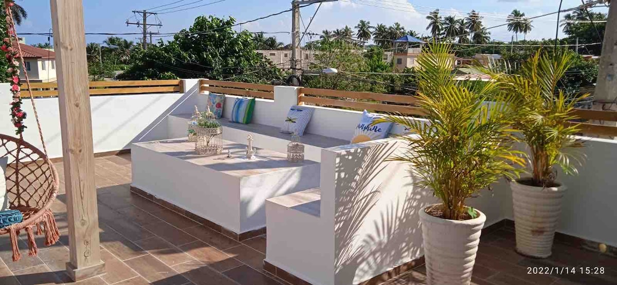 A shared terrace is furnished with white seating and vibrant cushions, surrounded by greenery. Potted plants add freshness to the space. Sunlight filtered through the trees casts soft shadows on the tiled floor, creating a serene outdoor area for relaxation.