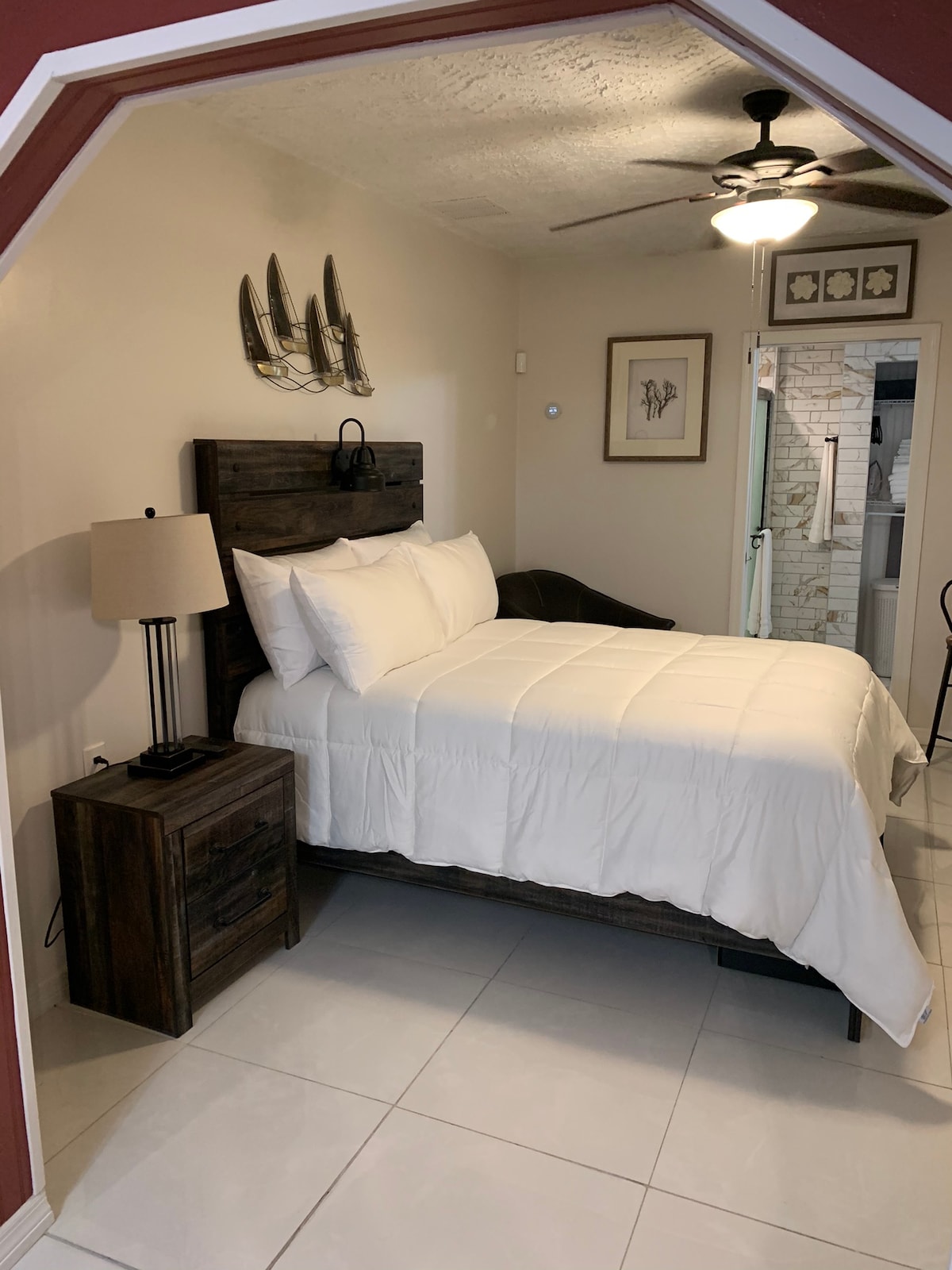 A cozy bedroom area is featured, showcasing a spacious bed dressed in white linens. A bedside table with a lamp is positioned alongside the bed. Natural light filters in, illuminating the room, which includes a glimpse of the bathroom area in the background.