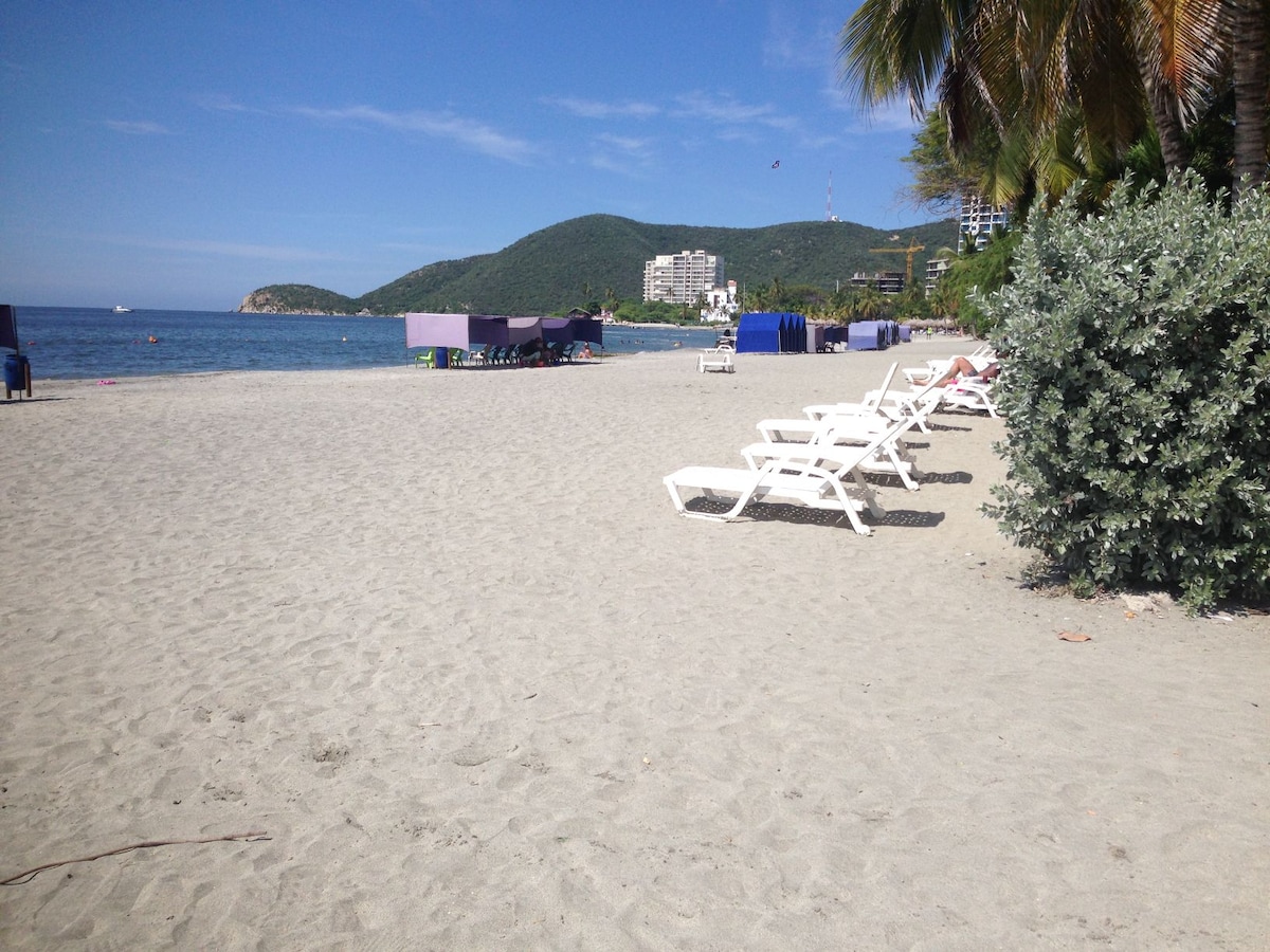 A sandy beach extends along the shoreline, with several white lounge chairs arranged in a row. The gentle waves of the sea are visible in the background, and hills covered with greenery rise behind the beach. Various umbrellas provide shaded areas in the distance.