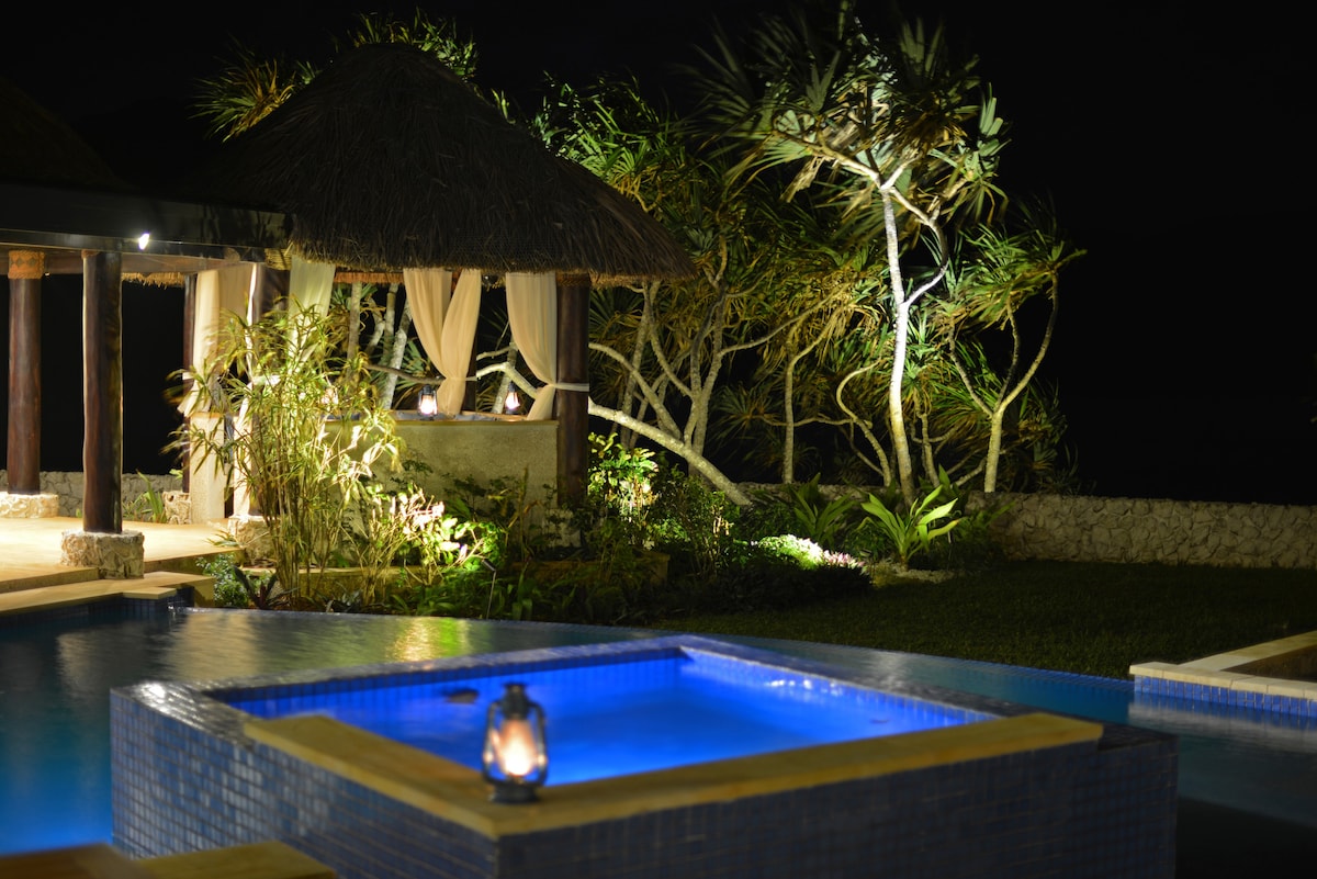 A tranquil evening scene is depicted, showcasing an illuminated spa tub surrounded by lush landscaping. Soft lighting highlights the nearby outdoor pavilion with draped fabric, enhancing the serene atmosphere against the night sky.
