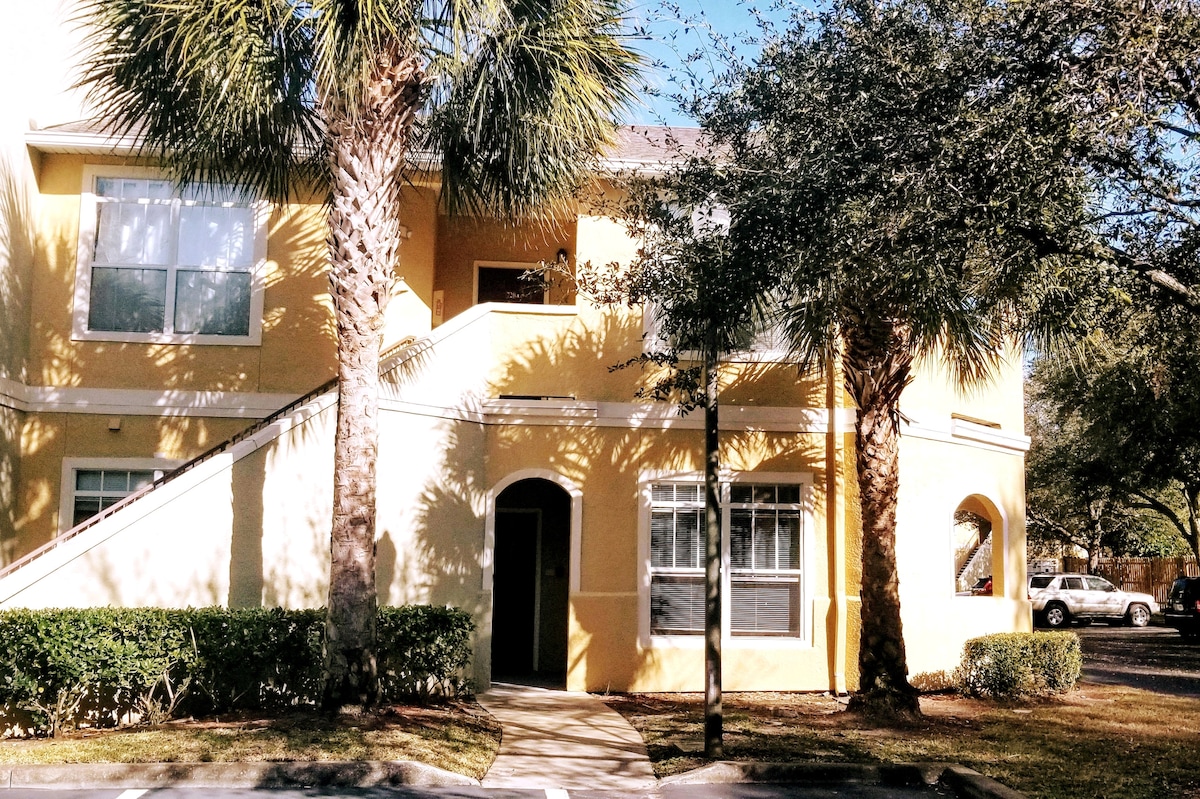 An exterior view of a yellow condo building is depicted, featuring palm trees and shrubs. A pathway leads to a private entrance, while large windows allow ample natural light. The surrounding landscaping creates a welcoming atmosphere in the gated community.