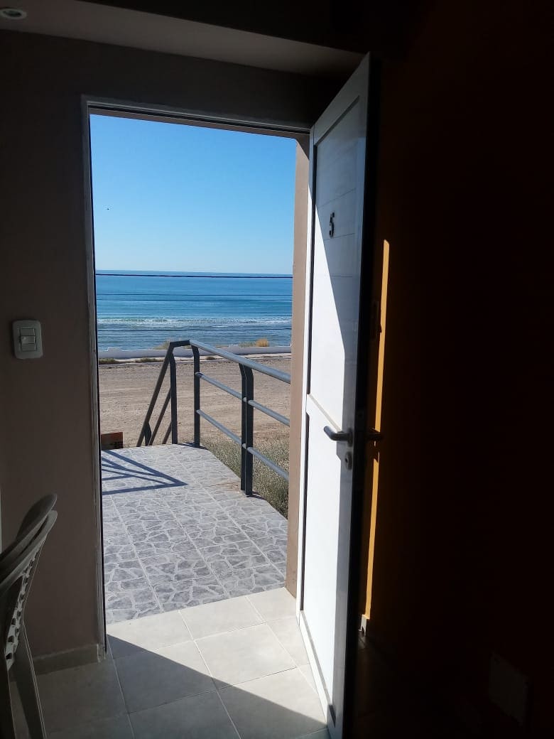 An open door reveals a private terrace overlooking the sea. A clear blue sky frames the coastal view, while a sandy beach stretches along the water's edge. The terrace features a railing, providing an unobstructed sight of the tranquil landscape.