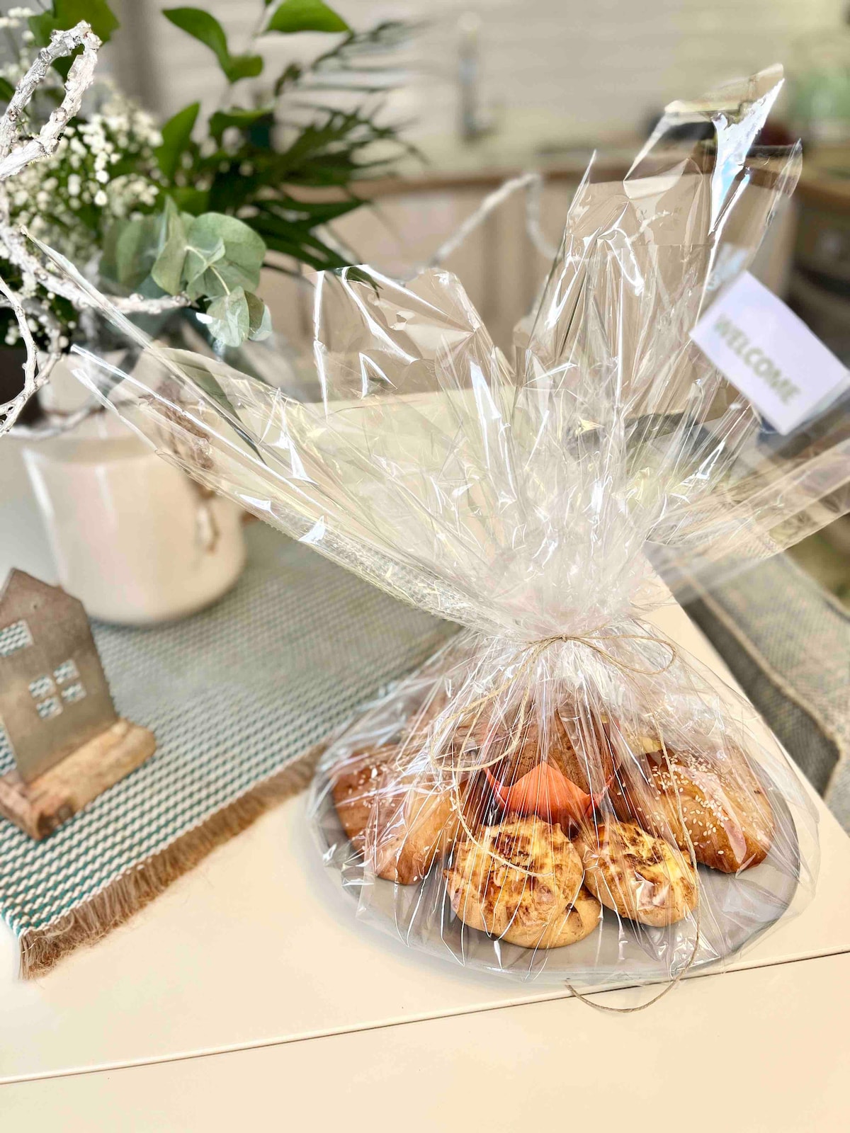 A plate of baked goods is presented in clear wrapping, showcasing an assortment of treats. The arrangement is placed on a textured surface, accompanied by a small decorative object and greenery. Soft light highlights the items, creating a welcoming presentation.