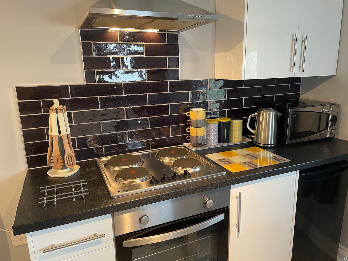 A modern kitchen area is presented with black tiled backsplash and light cabinetry. A stainless-steel oven and stovetop are visible, alongside a kettle and microwave. Colorful mugs are neatly arranged, with utensils displayed on a countertop, creating a functional cooking space.