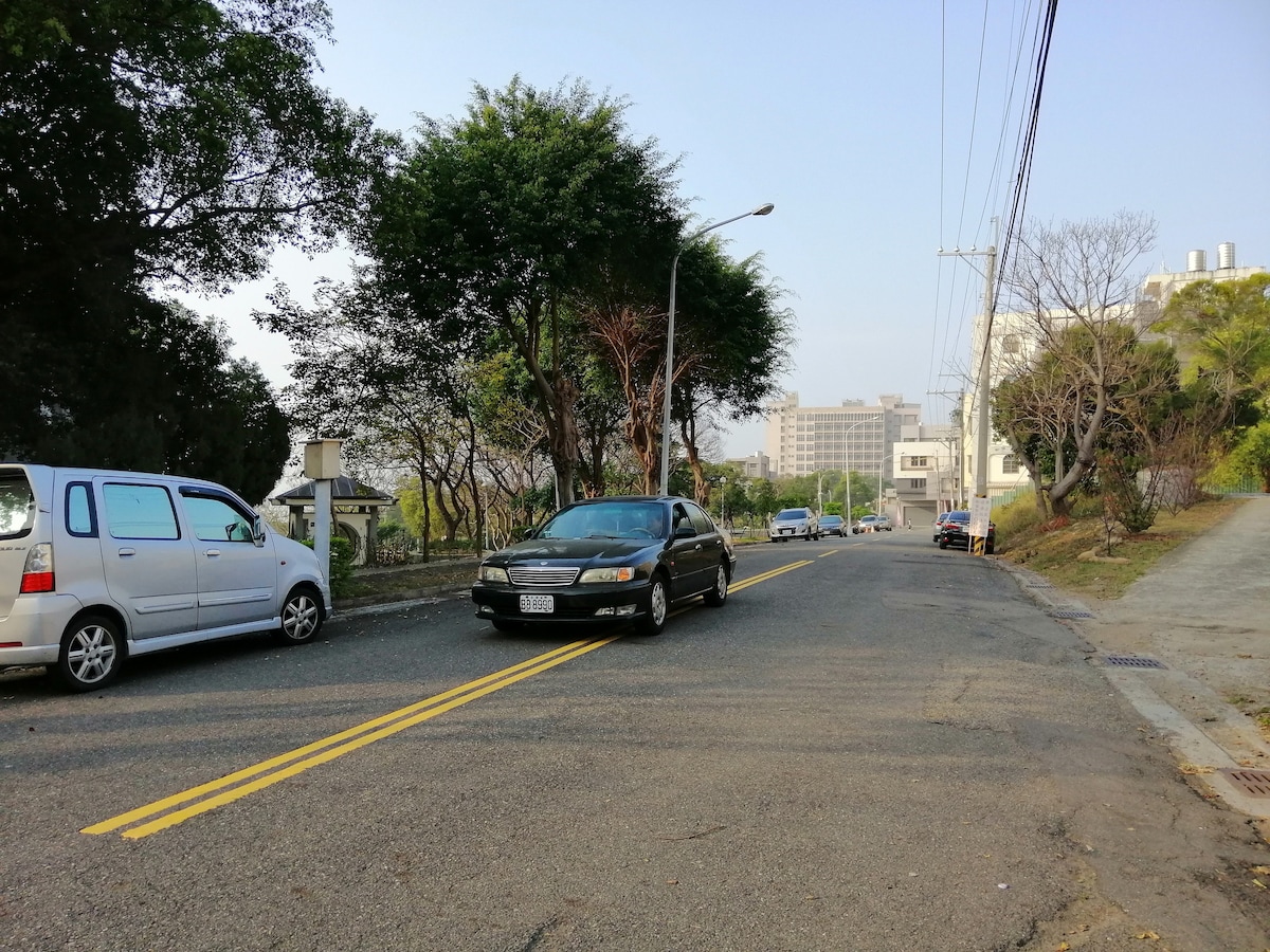 A quiet street scene shows a gently sloping road lined with trees on the left. Two cars are parked along the roadside, while one vehicle approaches from the opposite direction. In the background, buildings are visible against a clear sky.