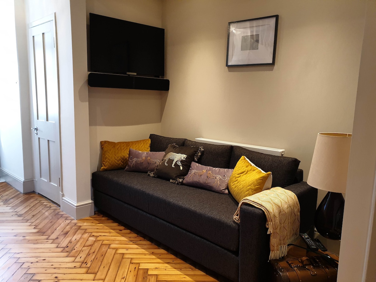 A dark grey sofa is positioned against a light wall, adorned with several decorative pillows in yellow and patterned fabric. A lamp with a beige shade is placed beside the sofa, and a flat-screen TV is mounted above the seating area. The floor features warm wooden herringbone patterns.