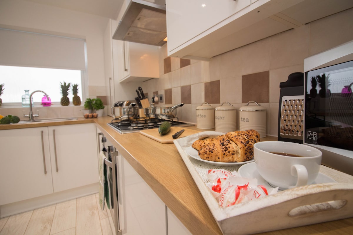 The kitchen features a modern design with wood accents and tiled walls. A white tray holds a cup of coffee, pastries, and sugar packets. Kitchen appliances, including a microwave and kettle, are visible, along with neatly arranged canisters and a sink by a window.