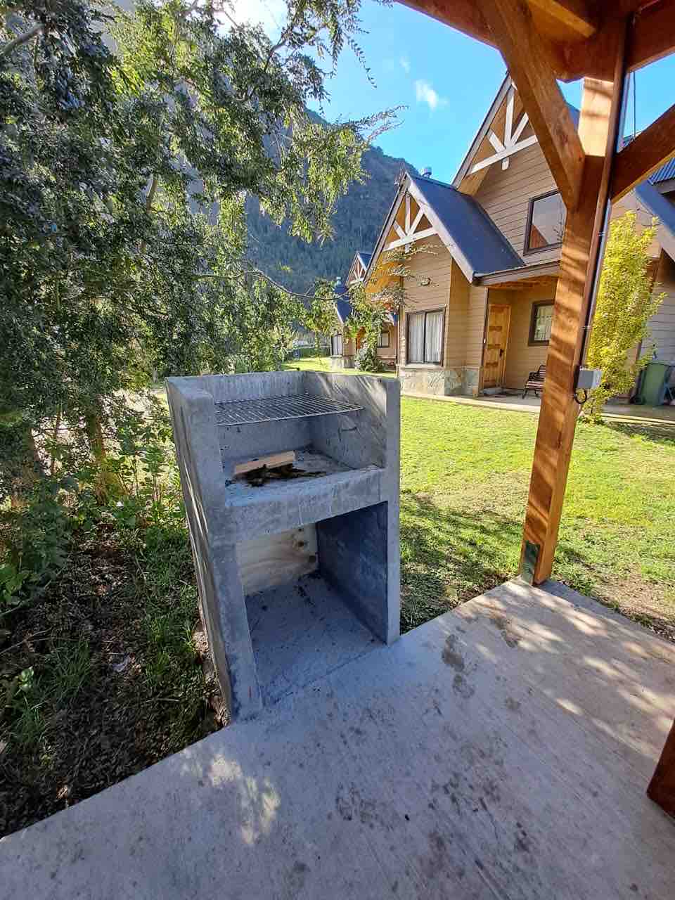 An outdoor grilling area is visible, featuring a built-in concrete barbecue. Lush greenery surrounds the space, with a glimpse of nearby wooden cabins in the background, and majestic mountains are seen rising above, completing the natural setting.