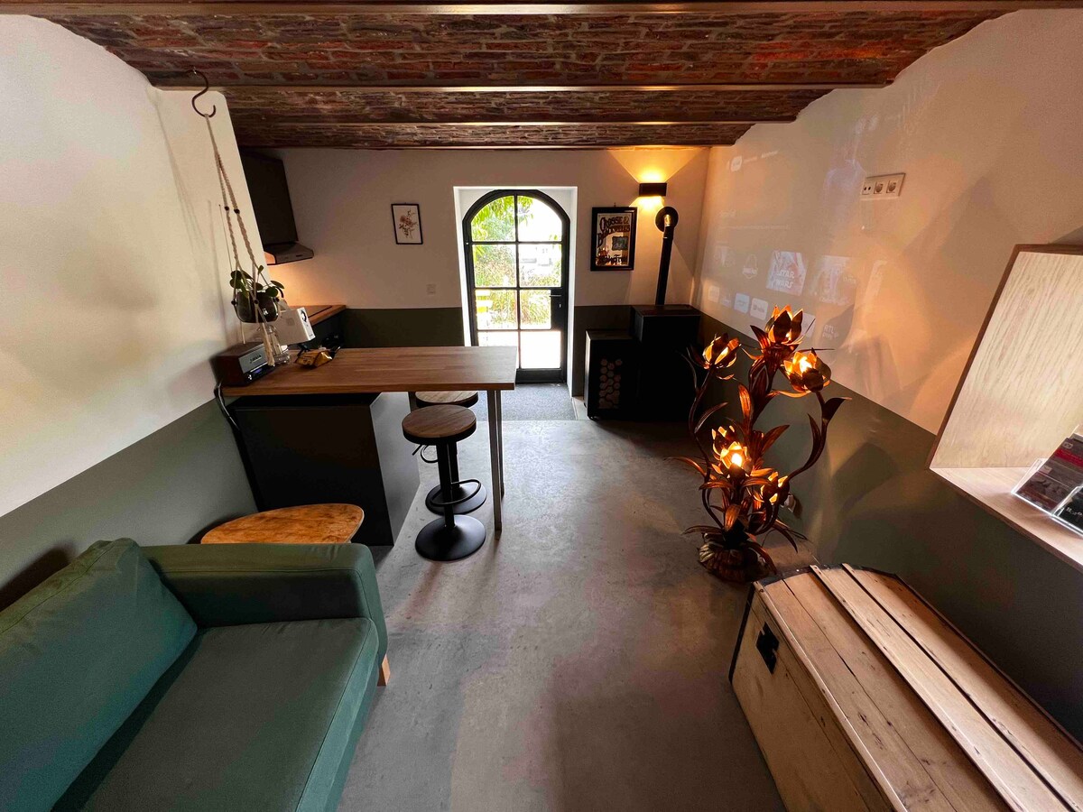 A cozy living area features a green sofa and a wooden dining table paired with a high stool. An eye-catching floor lamp with a floral design provides soft lighting, while a large arched window offers views of the outside garden.