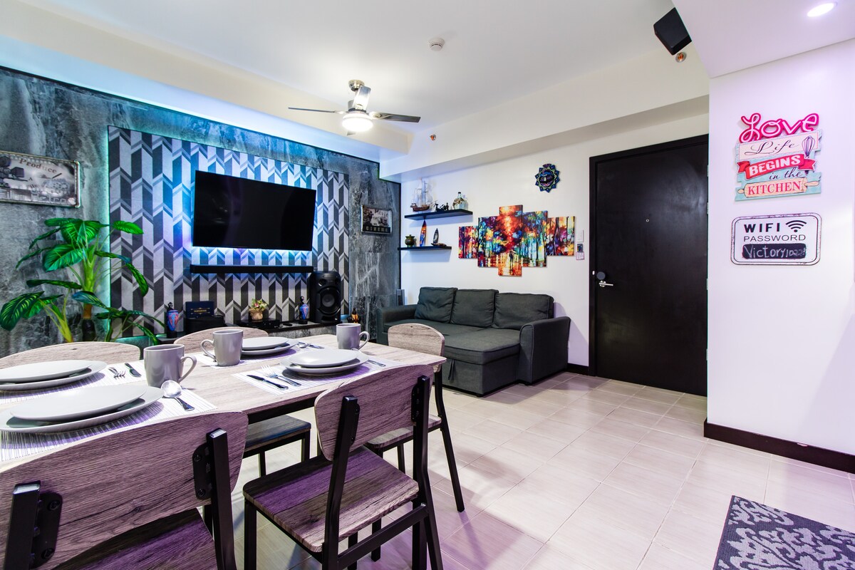 The living area features a modern dining table set for four, complemented by a cozy sofa and vibrant wall art. A sleek media wall holds a 55-inch television, with an illuminated background. Decorative elements, including plants, add a touch of greenery to the space.