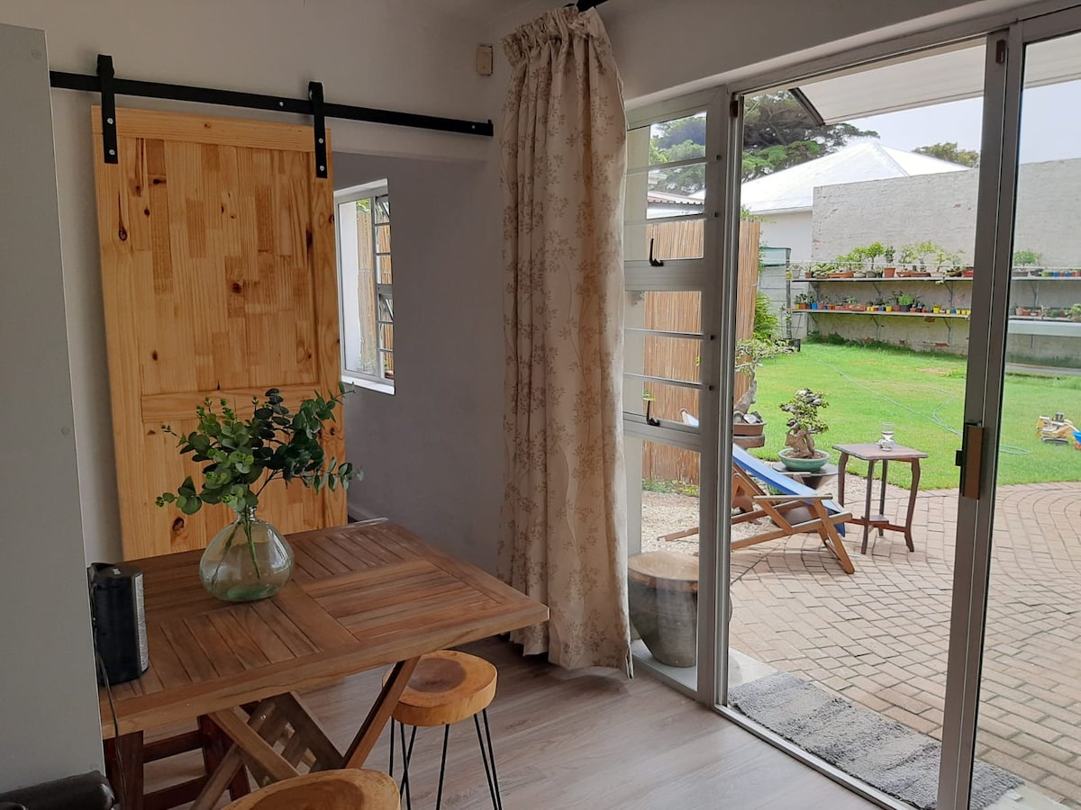 A dining area features a wooden table surrounded by three stools, complemented by a glass vase containing greenery. Natural light enters through large windows with sheer curtains, illuminating the space. An outdoor patio is visible beyond, showcasing a manicured lawn and potted plants.