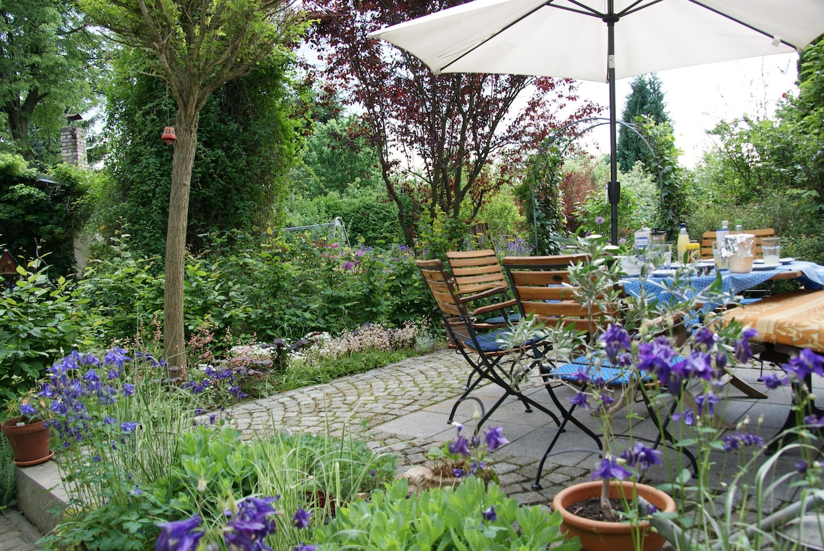 A serene garden space is presented, featuring a stone patio surrounded by lush greenery and vibrant purple flowers. A large umbrella provides shade over a wooden table and chairs, inviting relaxation amid the natural surroundings. The garden displays a variety of plants, enhancing the tranquil atmosphere.